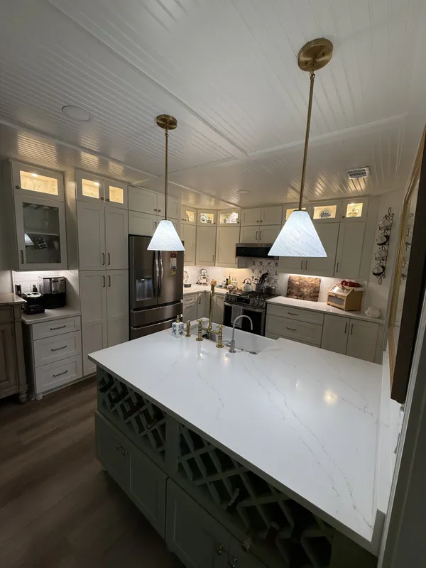 Finished kitchen with sage green island featuring built-in wine storage, white shaker perimeter cabinets, brushed brass fixtures, and beadboard ceiling.